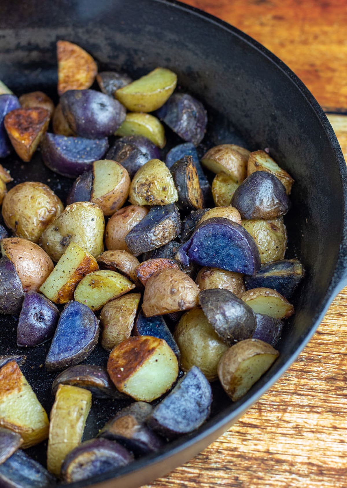 Multi-colored baby potatoes done being roasted in a large black cast iron skillet sitting on a wood background.