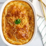 white casserole dish with dairy-free chicken pot pie in it, fresh out of the oven, sitting on white marble counterop, with a green and white striped towel and gold knife and fork beside it