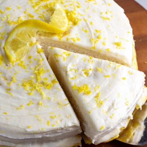dairy-free lemon cake on wooden cake stand with on piece cut and being pulled out with a cake server utensil