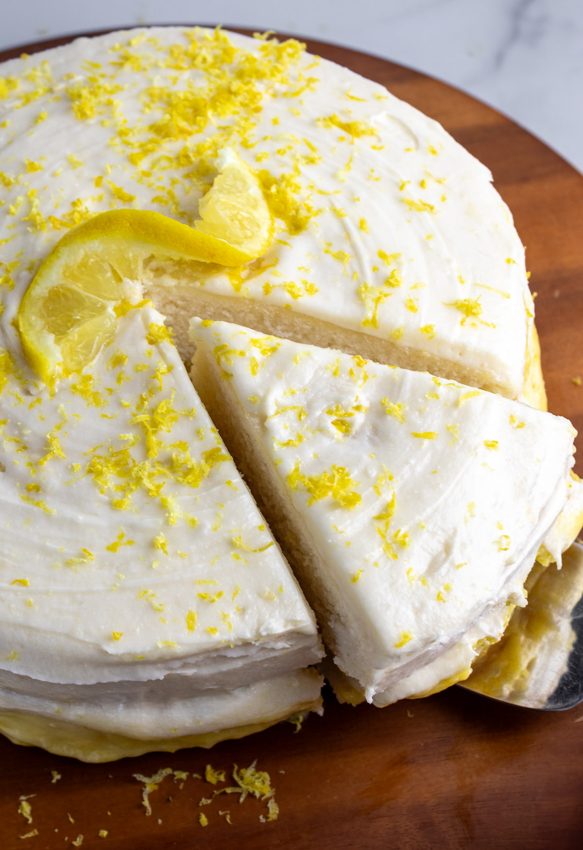 dairy-free lemon cake on wooden cake stand with on piece cut and being pulled out with a cake server utensil